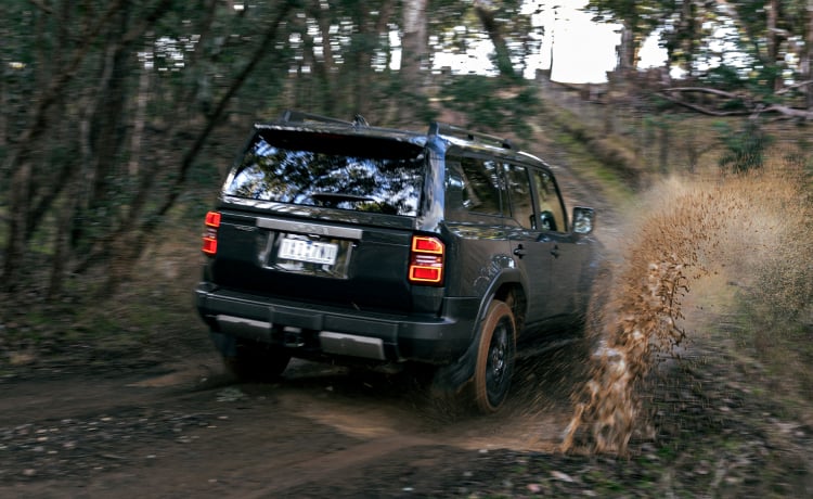 A Toyota LandCruiser Prado in Eclipse Black, driving down a dirt road in the bush, kicking up mud and water.