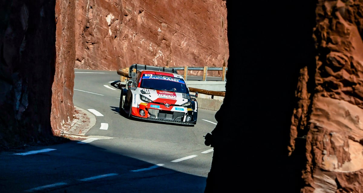 A Glacier White Toyota G R Yaris Rally 1, driving along a road that winds around a rocky red cliff. The vehicle is covered in sponsor livery.